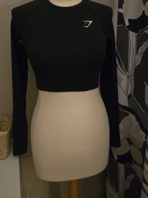 Svart crop top från Gymshark - Snygg svart långärmad crop top från Gymshark med rund halsringning och diskret logga på bröstet. Tillverkad i ett stretchigt funktionsmaterial som passar perfekt till träning eller gym. Tight passform och sportig look.