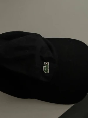 Svart keps från Lacoste - Snygg svart keps från Lacoste med den klassiska gröna krokodilloggan broderad framtill. Justerbart band bak i brunt läder med metallspänne och Lacoste-detaljer på insidan. Perfekt accessoar för en sportig och stilren look.