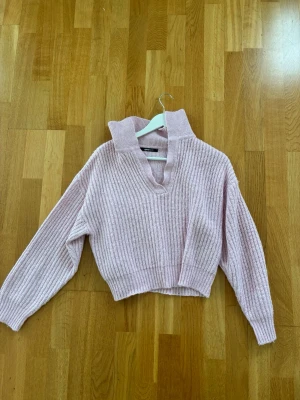 Ljusrosa stickad tröja med krage - Trendig ljusrosa stickad tröja med bred krage och v-ringning. Tröjan har ribbstickad struktur, långa ärmar och en något croppad passform. Perfekt för dig som gillar en avslappnad men ändå stilsäker look.