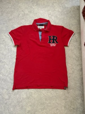 Röd pikétröja HR Twenty Seven - Snygg röd pikétröja från HR Twenty Seven med broderad logga och siffran 27 i marinblått på ryggen. Tröjan har klassisk krage, korta ärmar med vita detaljer och knappar vid halsen. Perfekt för en sportig och avslappnad stil.