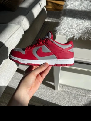 Nike Dunk Low röd/grå sneaker - Nike Dunk Low sneakers i rött och grått med klassisk Swoosh på sidan. Skon har en låg siluett, vita mellansulor och röda yttersulor. Tillverkad i läder med textildetaljer och röda skosnören. Perfekt för dig som gillar streetwear och vill sticka ut.