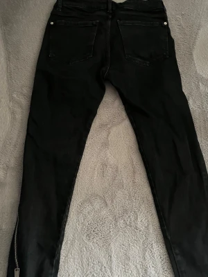 Svarta skinny jeans med dragkedja - Säljer ett par svarta skinny jeans med dragkedjedetalj längs hela sidan av benen. Jeansen har två bakfickor och är i stretchigt material för en tight passform. Perfekta för en edgy och trendig look.