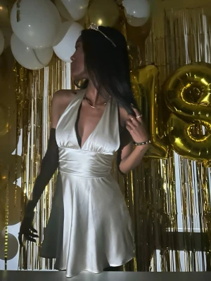 Champagnefärgad halterneck klänning - Snygg kort klänning i champagnefärgat satinliknande material med halterneck och djup v-ringning. Klänningen har öppen rygg och knytband i midjan för en smickrande passform. Perfekt för dig som vill sticka ut på festen!