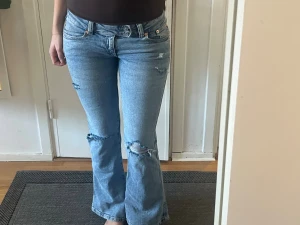 Ljusblå bootcut jeans med slitningar - Säljer ett par ljusblå bootcut jeans med slitna detaljer och hål vid knäna. Jeansen har klassisk femficksmodell, låg midja och är tillverkade i denim. Perfekta för en avslappnad och trendig look.