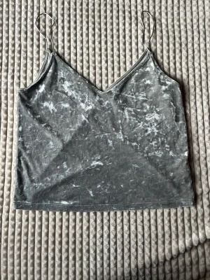 Grå sammetstopp med smala band - Säljer en grå croppad topp i mjuk sammet med tunna axelband och v-ringad hals. Toppen har en lätt glansig finish och är perfekt för dig som gillar en stilren men ändå trendig look.