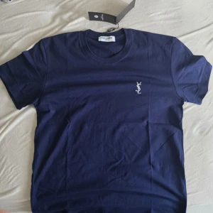 Mörkblå YSL t-shirt med logga - Snygg mörkblå t-shirt från Yves Saint Laurent med klassisk vit YSL-logga broderad på bröstet. T-shirten har rund hals och korta ärmar, perfekt för en clean och stilren look.