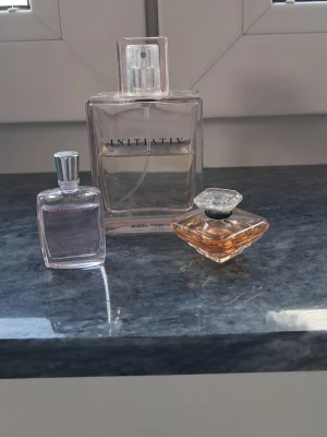 Tre parfymer från Initiativ & Lancôme - Säljer tre olika parfymer: en stor rektangulär flaska från Initiative med transparent design, en mindre rektangulär flaska från Lancôme med ljusrosa innehåll och silverfärgat lock samt en elegant, diamantformad flaska från Lancôme med bärnstensfärgad doft och genomskinligt lock.