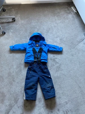 Mörkblå täckbyxor med hängslen - Säljer ett par mörkblå täckbyxor från Skifunk med justerbara svarta hängslen och dragkedja. Perfekta för vinterns alla äventyr. Materialet är slitstarkt och vattenavvisande, och byxorna har reflexdetaljer på benen.