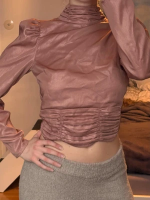 Rosa satinblus med öppen rygg - Snygg rosa blus i satin med hög krage, långa puffärmar och rynkade detaljer framtill. Baksidan har en stor öppen rygg och knyts med ett brett band i midjan. Perfekt för dig som vill sticka ut med en unik topp.