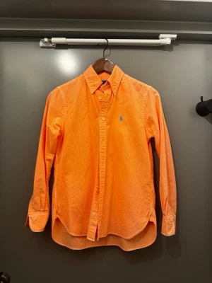  Polo Ralph Lauren skjorta -  Skjortan har klassisk krage, knappar framtill och det ikoniska blå broderade logotypen på bröstet. Perfekt för dig som vill sticka ut med färg och stil.