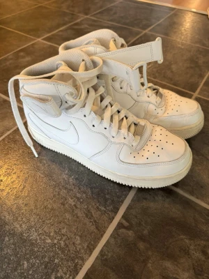 Nike Air Force 1 - Klassiska vita Nike Air Force 1 mids med kardborreband runt ankeln och perforerad tåbox. Skorna är tillverkade i läder med vit sula och ikonisk Swoosh på sidan. Perfekta för dig som gillar en clean och tidlös sneakerstil.