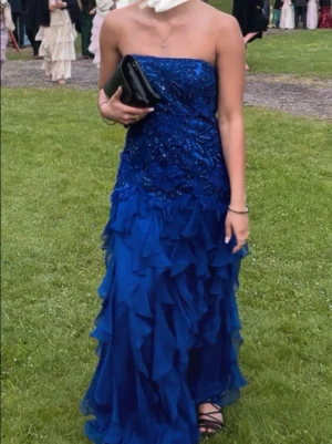 Blå axelbandslös balklänning - Säljer en fantastisk blå långklänning med axelbandslös design och vackra volangdetaljer. Överdelen har glittriga broderier och pärlor. Perfekt för bal eller festliga tillfällen. Klänningen har en figurnära passform upptill och faller snyggt i lager.