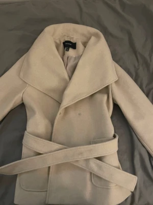 Beige kappa med skärp  - Stilren beige kappa  med bred krage och tillhörande skärp i midjan. Kappan har dubbelknäppning och två fickor framtill. Perfekt för dig som vill ha en klassisk och trendig look under kallare dagar.