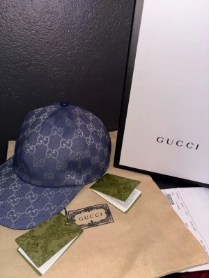 Blå denimkeps från Gucci - Säljer en blå keps från Gucci i denim med det klassiska GG-monogrammet över hela kepsen. Justerbar rem baktill i matchande material. Kepsen har vit insida och detaljer i läder. Perfekt för dig som vill ha en exklusiv och trendig accessoar.