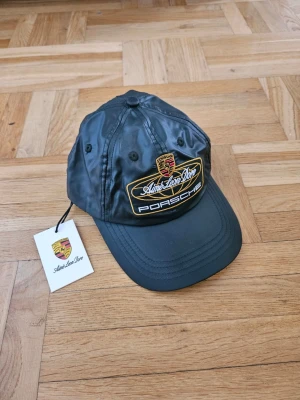 Aime Leon Dore x Porsche keps - Snygg mörkgrå keps från Aime Leon Dore i samarbete med Porsche. Kepsen har broderad Porsche-logga och gul/vit text framtill. Tillverkad i glansigt nylonmaterial med klassisk böjd skärm och justerbar passform. Perfekt för dig som gillar streetwear och bilar.