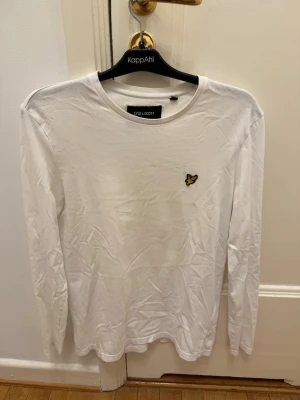 Vit långärmad t-shirt Lyle & Scott - Klassisk vit långärmad t-shirt från Lyle & Scott med rund hals och den ikoniska gula loggan broderad på bröstet. Tillverkad i mjuk bomull för en skön känsla och enkel stil som funkar till allt.