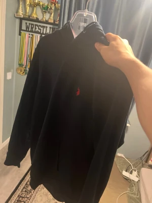 Svart hoodie från Polo Ralph Lauren - Snygg svart hoodie från Polo Ralph Lauren med klassisk röd logga broderad på bröstet. Hoodien har huva med dragsko och en avslappnad passform. Perfekt för en stilren och bekväm look.