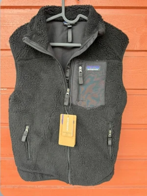 Patagonia väst  - Mysig svart fleeceväst från Patagonia med dragkedja framtill, tre fickor med dragkedja och en bröstficka i nylon. Ej autentisk. Perfekt för lager-på-lager och outdoor vibes. Klassisk hög krage och mjukt teddy-material.