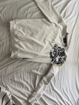 Philipp Plein long sleeve small - Vit sweatshirt från Philipp Plein med stor broderad lejon-design i svart och guld på bröstet och axeln. Tröjan har rund halsringning, ribbade muddar och är tillverkad i mjukt bomullsmaterial. Perfekt för dig som gillar statement-plagg och exklusiva detaljer.