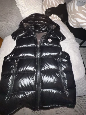 Moncler väst  - Snygg svart dunväst från Moncler med glansig finish och huva. Västen har dragkedja framtill, två stora fickor och Moncler-logga på bröstet. Insidan har ett unikt serietidningsmönster och märkets etikett. Perfekt för lager på lager och streetwear. Kommer med NFC tag 