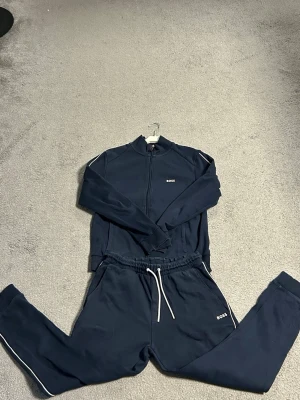 Marinblått loungewear-set från BOSS - Mysigt marinblått loungewear-set från BOSS med dragkedja på tröjan och vita snören i midjan på byxorna. Setet har diskreta BOSS-loggor på både överdel och byxa samt vita detaljer längs bensluten. Perfekt för chill dagar hemma.