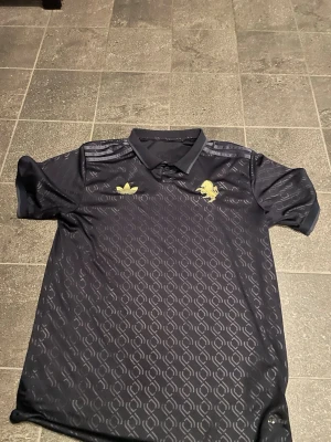 Svart Adidas sporttröja med mönster - Säljer en svart sporttröja från Adidas med diskret mönster och guldfärgade detaljer. Tröjan är från fotbollsklubben Juventus och detta var deras tredje ställ några år tillbaka. Tröjan har krage, korta ärmar och klassiska Adidas-ränder på axlarna. Materialet är troligtvis polyester, perfekt för träning eller sport. Fungerar även som en vanlig tröja till tex en chill utekväll.