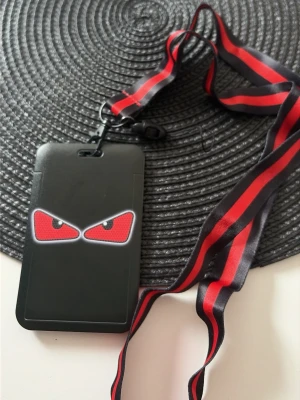 Svart korthållare med röd lanyard - Cool svart korthållare med röda ögonmotiv och tillhörande lanyard i svart och rött. Perfekt för att bära ID-kort eller passerkort runt halsen. Korthållaren har ett skyddande fack och snodden har ett smidigt clips.