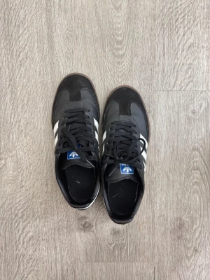 Adidas Samba svarta sneakers - Svarta Adidas Samba sneakers med klassiska vita ränder på sidorna och brun gummisula. Skorna har låg profil och är tillverkade i läder och mocka. Logotypen och texten 'SAMBA' i guld på sidan ger en ikonisk look. Perfekta för dig som gillar retrostil.