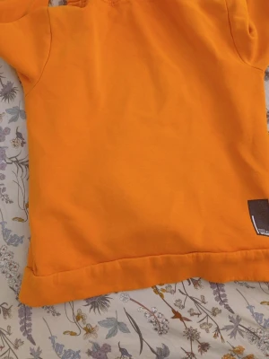Orange hoodie med tryck - Snygg orange hoodie från Name It med svart tryck på bröstet och stor känguruficka framtill. Hoodien har en klassisk huva och är gjord i mjukt bomullsmaterial. Perfekt för dig som gillar färg och vill sticka ut lite extra.