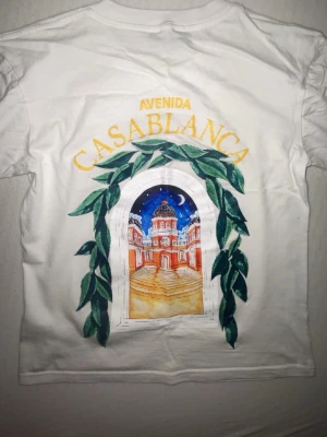 Vit t-shirt från Casablanca - Vit t-shirt från Casablanca med färgglatt tryck på ryggen som föreställer en byggnad och gröna blad, samt texten 'Avenida Casablanca'. Klassisk rund hals och korta ärmar. Perfekt för dig som gillar unika prints och streetstyle.
