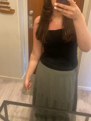 Grön långkjol  - Säljer en grön långkjol i storlek M. Kjolen har en smockad midja och faller snyggt i flera lager för en avslappnad look. Perfekt att styla med ett linne eller crop top för en chill vibe.