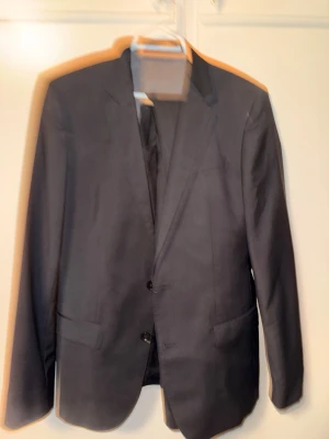 Svart klassisk kostymkavaj - Snygg svart kavaj med två knappar och klassisk passform från Hugo boss. Kavajen har släta slag, innerfoder och raka fickor framtill. Perfekt till kostymbyxor eller för att dressa upp en outfit. Storlek M men passar L