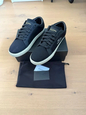 Svarta Saint Laurent sneakers - Svarta sneakers från Saint Laurent med vit sula och broderad logga på sidan. Skorna har klassisk låg modell, svarta skosnören och detaljer i textil och läder. Perfekta för en stilren och trendig look.