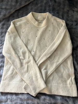 Vit stickad tröja med pärlor - Säljer en vit stickad tröja från H&M med pärldetaljer framtill, baktill och på ärmarna. Tröjan har rund halsringning, långa ärmar och breda ribbade muddar vid ärmslut och nederkant. Lite nopprig men inget som märks av! 