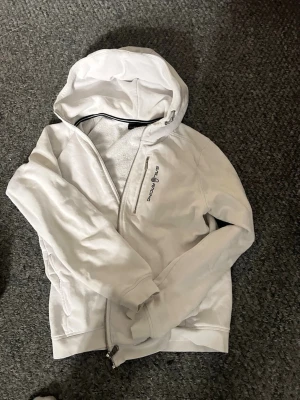 Vit hoodie från Sail Racing - Snygg vit hoodie från Sail Racing med dragkedja och broderad logga på bröstet. Hoodien har en stor huva, ribbade muddar och mjukt material på insidan. Perfekt för chill dagar eller när du vill ha en clean look. Säljes i befintligt skick