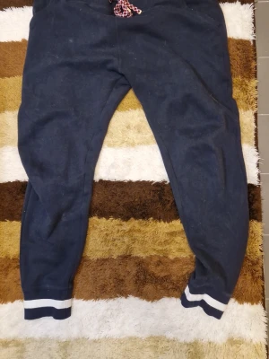 Mörkblå mjukisbyxor Tommy Hilfiger - Mörkblå mjukisbyxor från Tommy Hilfiger med vita och röda detaljer på snöret i midjan. Byxorna har muddar med vita ränder vid bensluten och är tillverkade i mjukt material för maximal komfort. Perfekta för chill dagar eller när du vill vara extra bekväm.