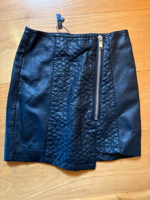 Svart quiltad skinnkjol med dragkedja - Snygg svart kortkjol i skinnimitation med quiltade detaljer framtill och en sned dragkedja som ger en edgy look. Kjolen har en tight passform och är perfekt för att lyfta vilken outfit som helst.