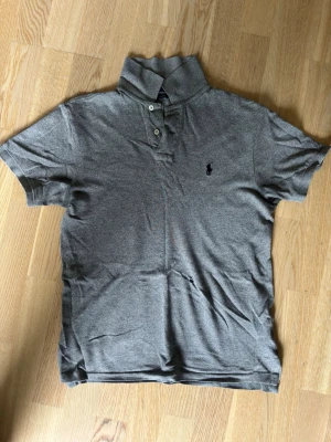 Grå polo t-shirt från Ralph Lauren - Säljer en grå polo t-shirt från Ralph Lauren med klassisk krage och tre knappar framtill. T-shirten har korta ärmar och en diskret broderad logga på bröstet. Perfekt för dig som vill ha en stilren och enkel look. Ett litet hål vid magen därav priset.