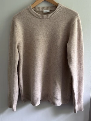 Beige stickad tröja i ull och kashmir strl M - Klassisk beige stickad tröja från ARKET i en mjuk mix av ull och kashmir. Tröjan har rund halsringning, långa ärmar och ribbade muddar vid ärmslut och nederkant. Perfekt för lager på lager och enkel att matcha. Endast använd ett fåtal gånger.