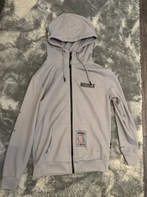 Ljusgrå hoodie från Hoodrich Storlek S - Snygg ljusgrå hoodie från Hoodrich med dragkedja framtill, stor huva och tryck både på bröstet, ryggen och ärmen. Hoodien har fickor fram och coola detaljer som text och logga i svart och rött. Storlek S