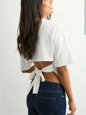 Vit topp med knyt i ryggen - Snygg vit topp med kort ärm och öppen rygg som knyts baktill. T-shirten har en croppad passform och är tillverkad i mjuk bomull. Perfekt för en trendig och avslappnad look.