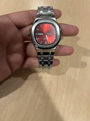 Röd Seiko herrklocka i stål - Stilren Seiko herrklocka med röd urtavla och dag- och datumvisning. Klockan har ett klassiskt armband i silverfärgat stål och markerade index. Perfekt accessoar för dig som vill sticka ut med en färgstark detalj.