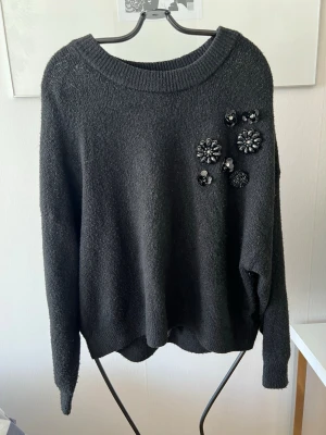 Svart stickad tröja med blommor - Mysig svart stickad tröja med rund halsringning och dekorativa blommor i paljetter och pärlor på ena axeln. Tröjan har en avslappnad passform och långa ärmar, perfekt för att ge outfiten lite extra edge.