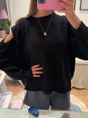Svart stickad tröja med cut out-detalj - Trendig svart stickad tröja med cut out-detalj på båda axlarna och avslappnad passform. Den är lite nopprig men går enkelt att fixa med noppermaskin 