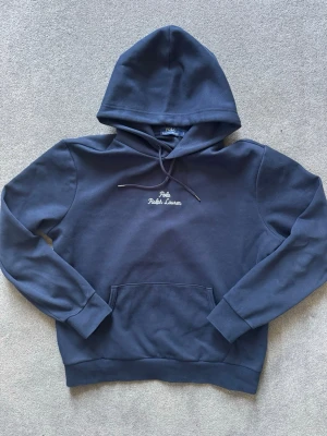 Mörkblå/ marin  hoodie från Polo Ralph Lauren - Mörkblå/ marin  hoodie från Polo Ralph Lauren med broderad logga på bröstet och liten brodyr på ärmen. Klassisk känguruficka framtill, dragsko i huvan och mjukt bomullsmaterial. Perfekt för en chill och stilren look. (Pris kan diskuteras)