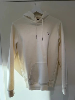Vit hoodie från GANT med logga - Snygg vit hoodie från GANT med liten broderad logga på bröstet. Klassisk modell med huva och dragsnören, samt en stor magficka framtill. Tillverkad i mjukt bomullsmaterial och har ribbade muddar vid ärmslut och nederkant.
