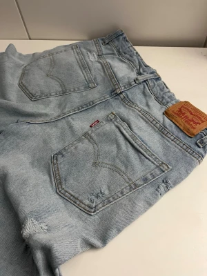 Ljusblå slitna Levi's jeans - Ljusblå jeans från Levi's med klassisk femficksdesign och slitna detaljer framtill och baktill. Jeansen har raka ben och är tillverkade i denim. Perfekta för en avslappnad och trendig look.