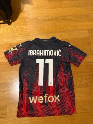 AC Milan Ibrahimović matchtröja - Snygg AC Milan matchtröja med Ibrahimović och nummer 11 på ryggen. Tröjan är svart med röda grafiska detaljer, korta ärmar och guldiga loggor från Puma och Emirates. Tillverkad i ett lätt och ventilerande material, perfekt för fotboll eller träning.