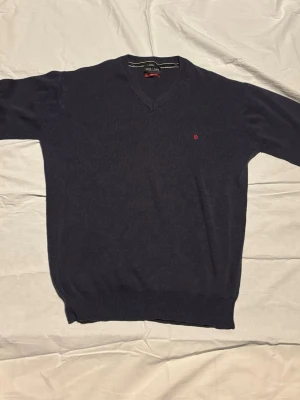 Mörkblå stickad tröja från Polo Ralph Lauren - Mörkblå stickad tröja från Polo Ralph Lauren med V-ringning och diskret röd logga broderad på bröstet. Tröjan har långa ärmar och ribbade muddar vid ärmslut och nederkant. Perfekt för en stilren och klassisk look.