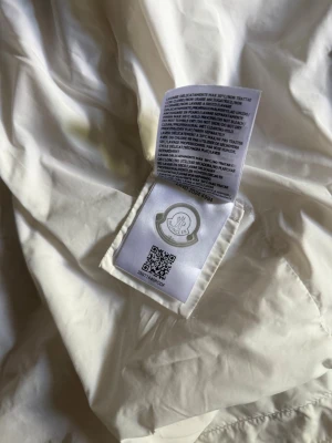 Vit dunjacka från Moncler - Snygg vit dunjacka från Moncler med klassisk logotyp på insidan. Jackan är tillverkad i ett glansigt material och har en lätt puffig form som ger en trendig siluett. Perfekt för kalla vinterdagar och riktigt varm tack vare dunfyllningen.